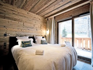 Ein schlicht eingerichtetes Schlafzimmer im Chalet-Stil mit einem großen Bett, einem Fenster mit Vorhängen und Blick auf eine Holzterrasse.
