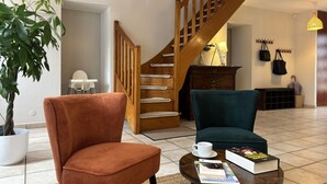 A living room with a wooden staircase, a potted plant, a red chair, a teal armchair, and a coffee table with books and a cup.