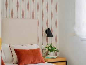 Ein Schlafzimmer mit einem Bett, Kissen, einem Nachttisch mit Lampe, einer Blumenvase und floraler Tapete.