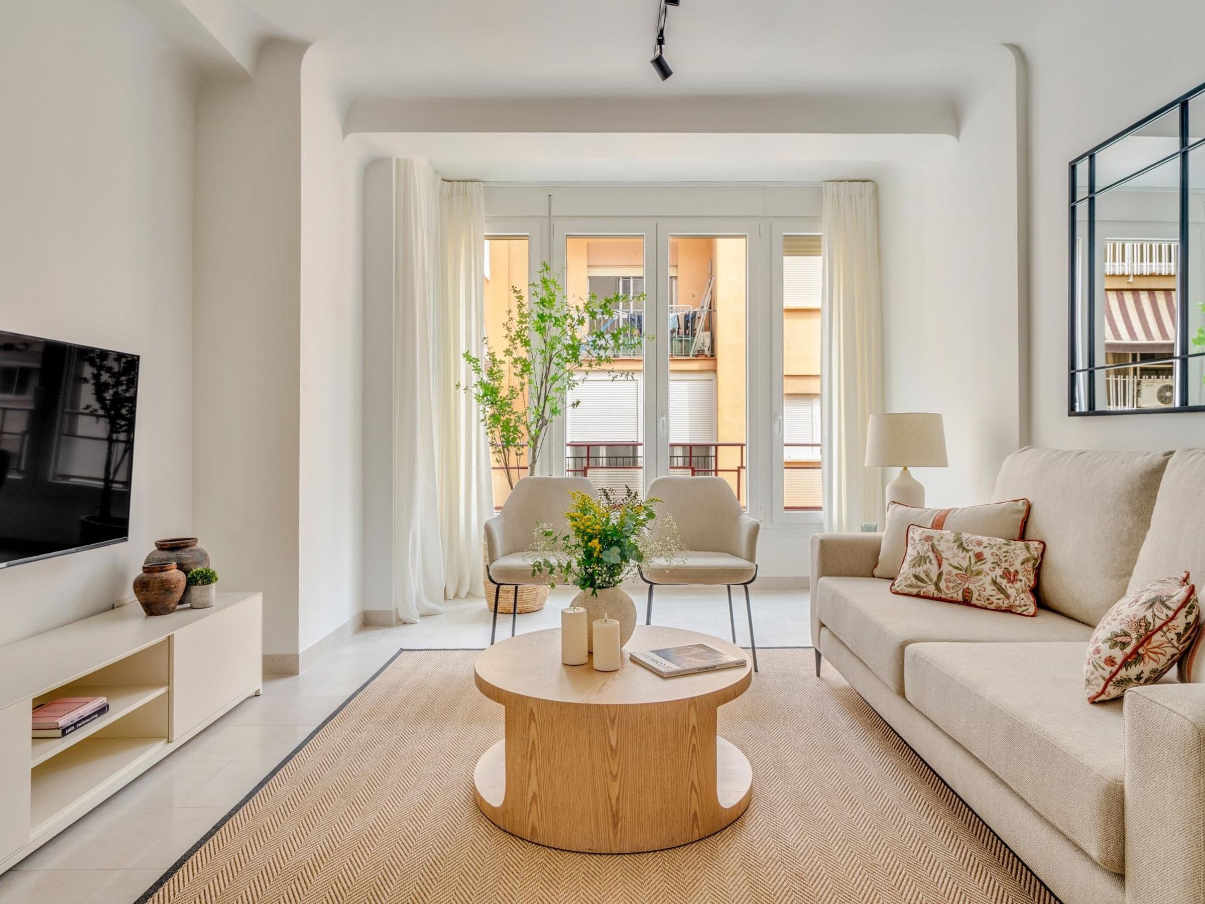 A modern living room with a flat-screen TV, a beige sofa, a round coffee table, and a view of a balcony with plants.