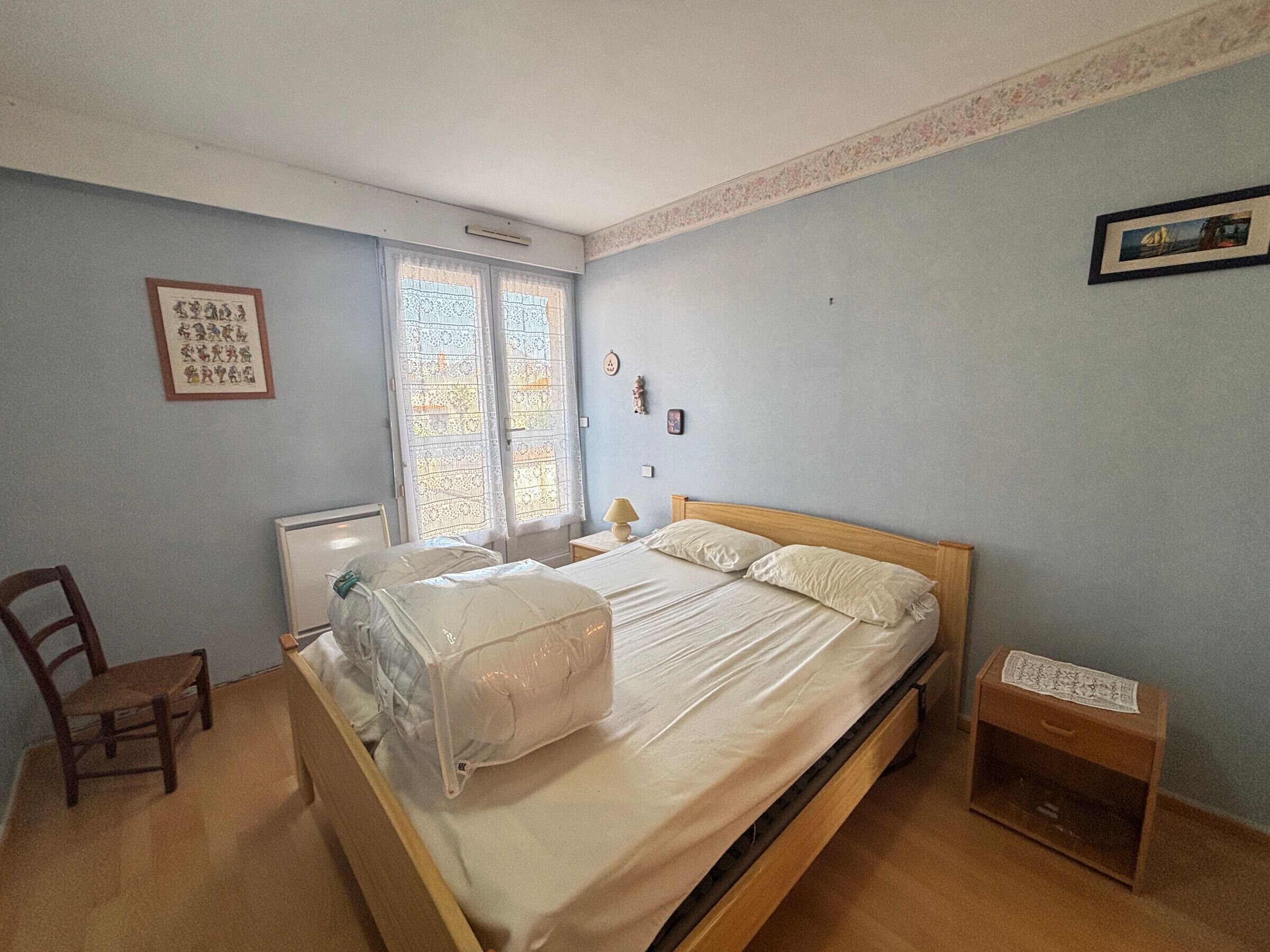 A bedroom with a single bed, a chair, a bedside table, a lamp, and a window with lace curtains.