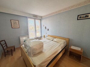 Ein Schlafzimmer mit einem Einzelbett, einem Sessel, einem Nachttisch, einer Lampe und einem Fenster mit Spitzenvorhängen.
