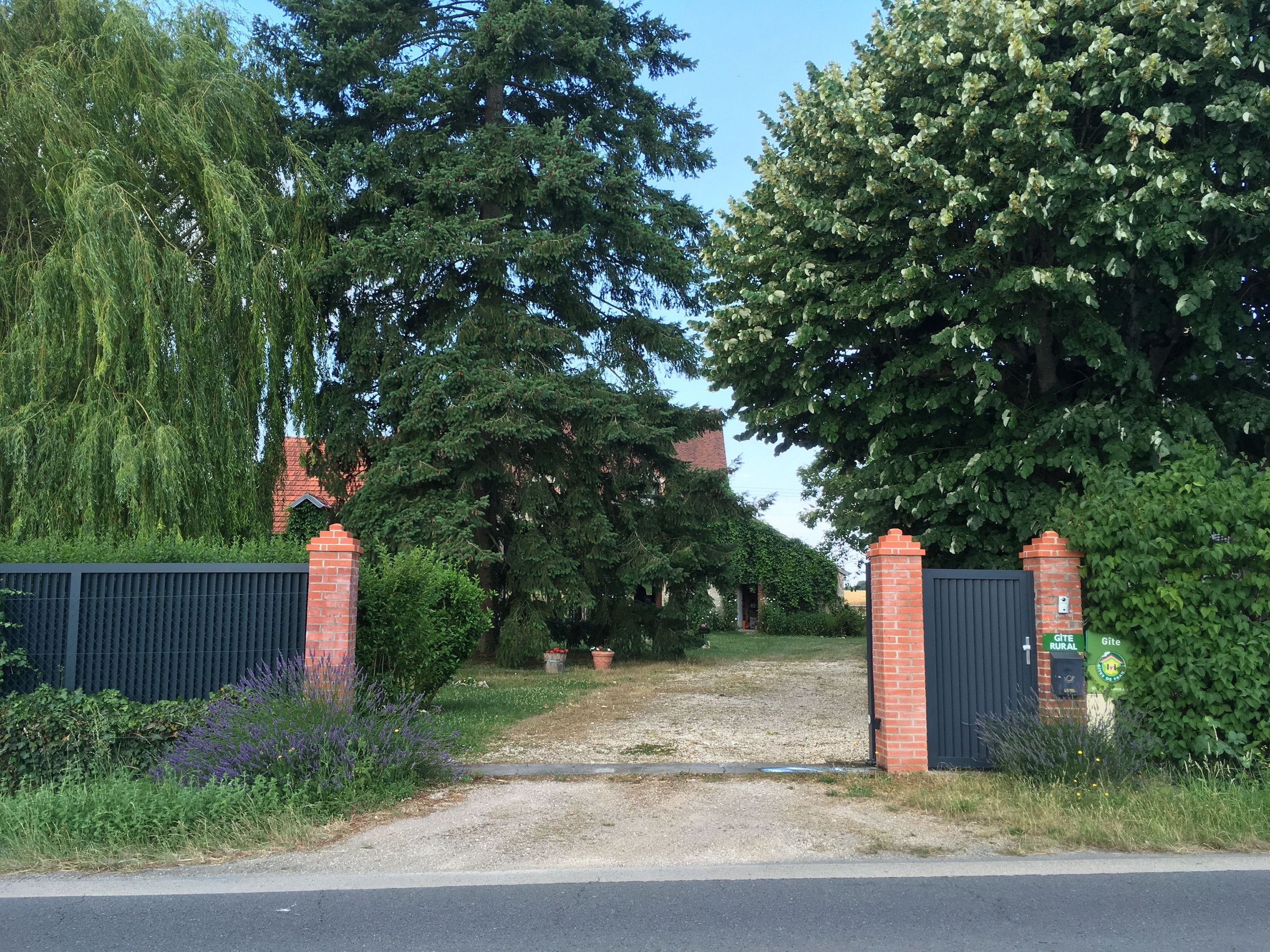 Een toegangshek met een grindpad dat naar een gebouw leidt, omgeven door weelderige begroeiing en bomen.