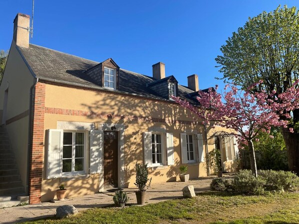 A two-story house with a brick facade, white shutters, and a chimney. There is a garden with a flowering tree and potted plants.