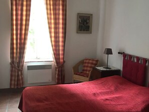 A room with a bed, a chair, a small table with a lamp, and a window with curtains.