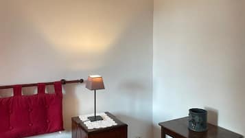 A room with a bed, a bedside table with a lamp, a small table with a decorative item, and a wall with a curtain.