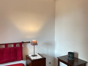 A room with a bed, a bedside table with a lamp, a small table with a decorative item, and a wall with a curtain.