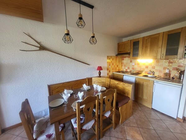 Zona pranzo con tavolo e sedie in legno, cucina con pensili in legno, zoccolo piastrellato e illuminazione a sospensione.