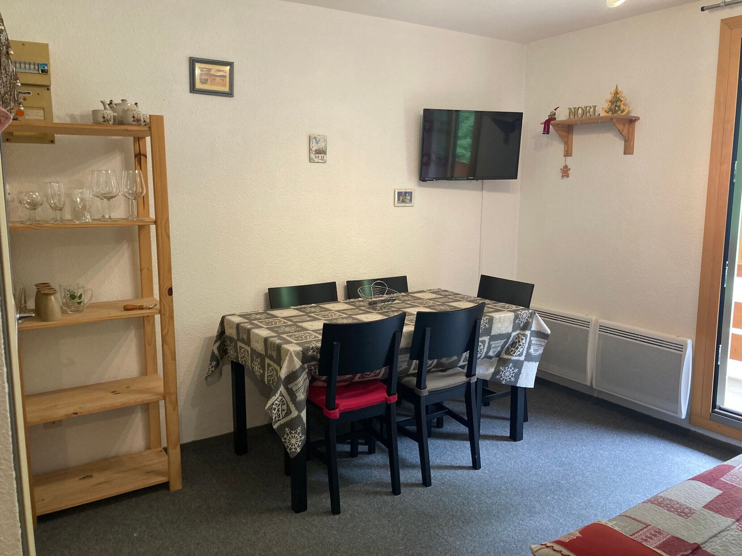 A dining area with a table set for four, surrounded by chairs, a wooden shelf with glassware, and a television mounted on the wall.