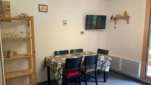 A dining area with a table set for four, surrounded by chairs, a wooden shelf with glassware, and a television mounted on the wall. - Apartment 4 guests, slope view - Le Pleynet (Le Pleynet)