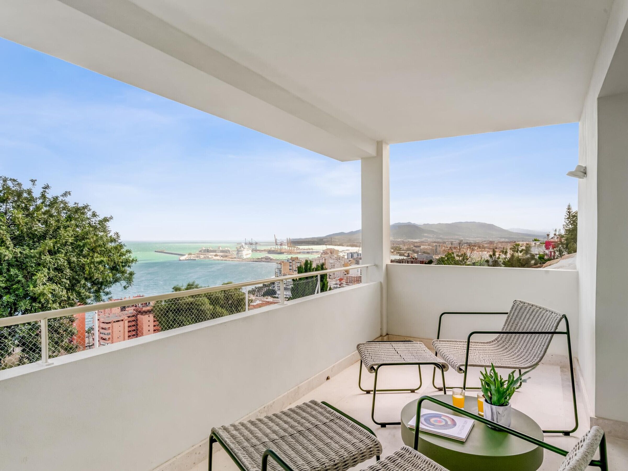 Een balkon met uitzicht op zee en stadsbeeld, voorzien van rieten stoelen en een tafel.