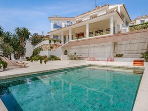 Ein moderner Poolbereich mit einem kühlen, blauen Pool, einer überdachten Sitzgelegenheit und einem mehrstöckigen Gebäude mit weißer Fassade und roten Akzenten.