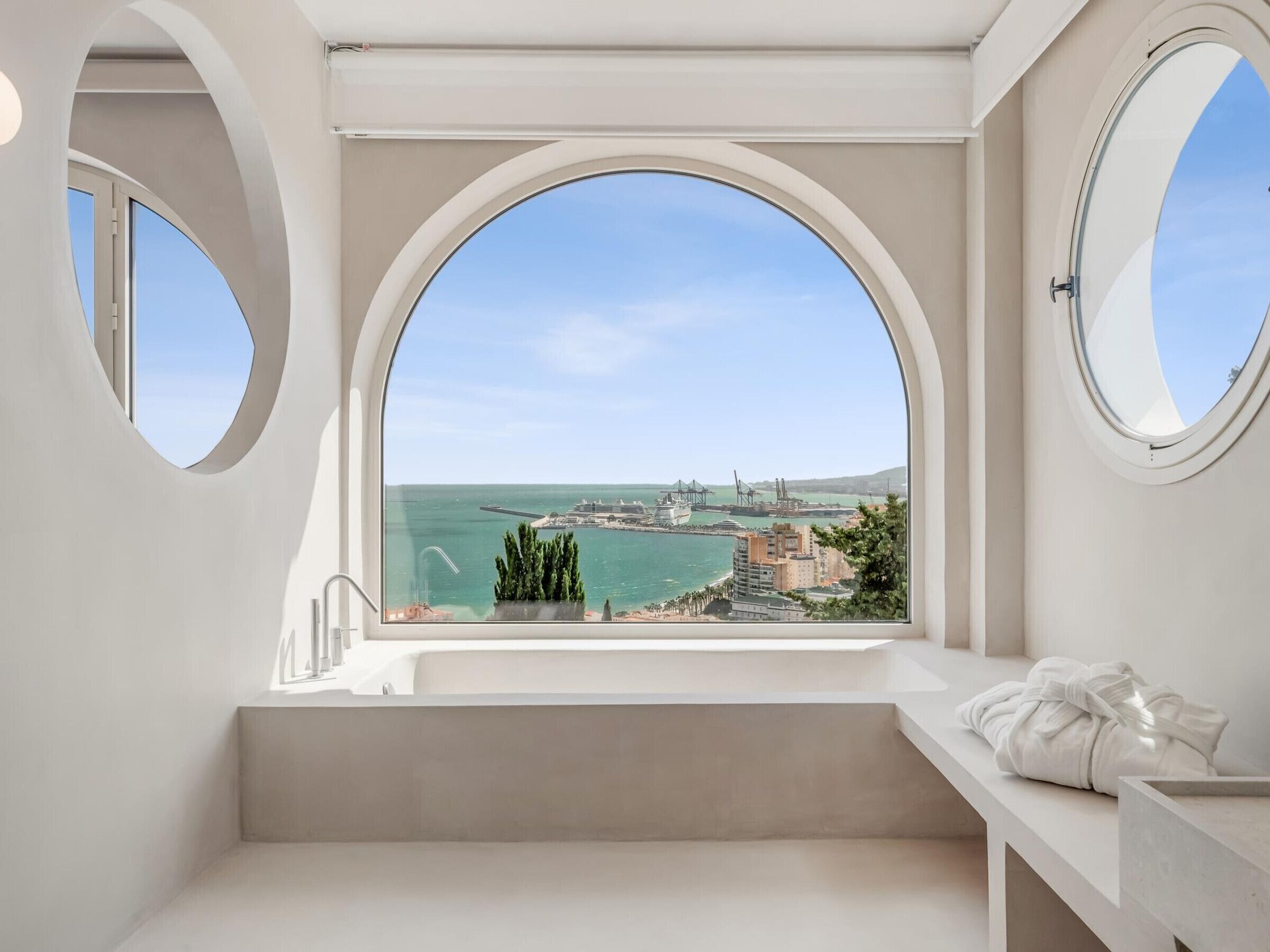 A bathroom with a large arched window overlooking a coastal cityscape.