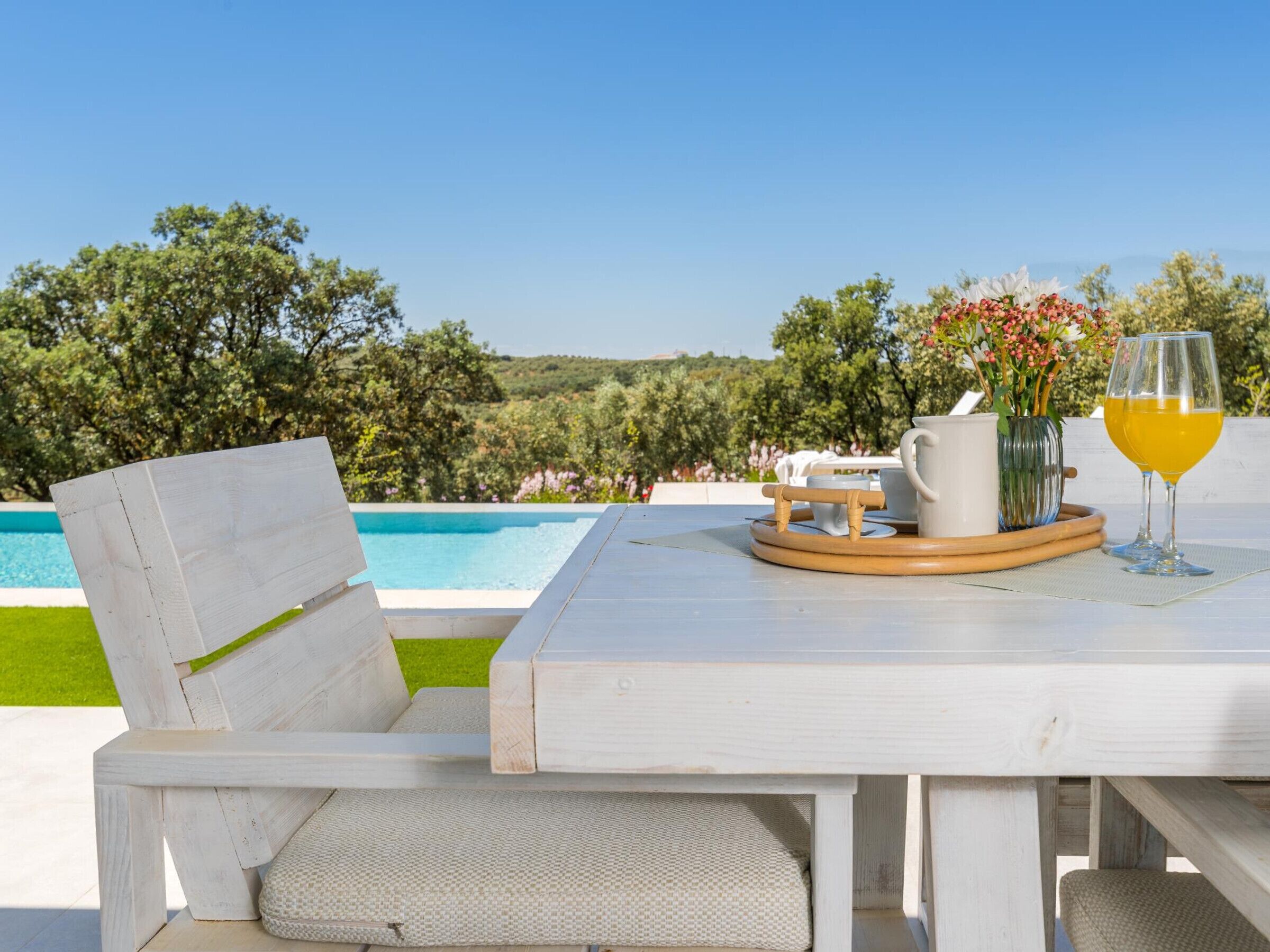 Une table blanche pour l’extérieur, avec une chaise, une carafe, un verre de jus d’orange et un vase rempli de fleurs.