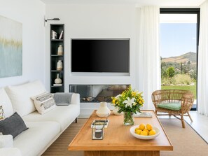 A modern living room with a white sofa, a flat-screen TV, a fireplace, and a view of the outdoors.