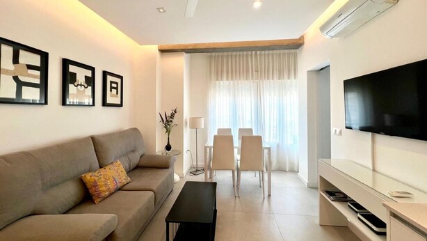 A modern living room with a sofa, a coffee table, a dining area with chairs, a television, and an air conditioning unit. - Central apartment in Málaga - 2 guests (Málaga)