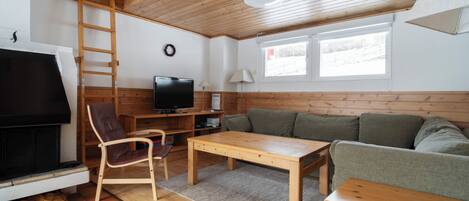 Ein gemütliches Wohnzimmer mit Holzbalkendecke, einem Fernseher, einer Couch, einem Tisch und einem Sessel.