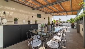 Eine Terrasse im Freien mit einem Glastisch und Stühlen, umgeben von Blumentöpfen und einem schwarzen Schrank.