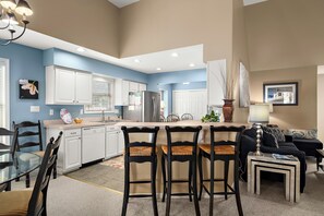 A modern kitchen with white cabinets, a central island with bar stools, and a dining area with a black sofa and a small table. - Pond Front Home with Magnificent Sunset views from the gorgeous Screened porch! (Lewes)