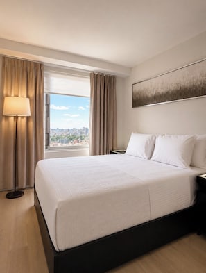 Et hotelværelse med en stor seng, et natbord, en lampe og udsigt til byens skyline gennem vinduet.