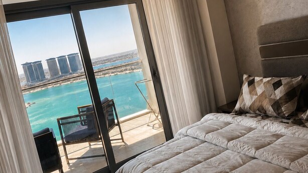A bedroom with a large bed, a view of a body of water, and a balcony with a chair. - Naya Suites; Luxury 2BR Panoramic Sea & Lagoon View, Beach Towers (Marsa Matrouh)