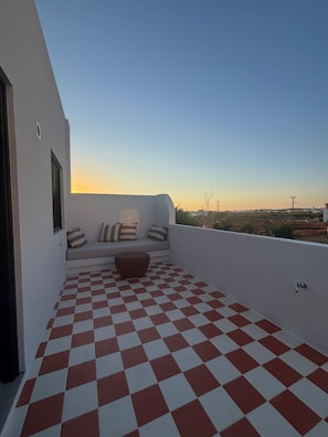 A balcony with a checkered floor, a seating area with cushions, and a view of the landscape.