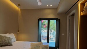 A hotel room with a bed, a ceiling fan, a window with a view of the outdoors, and a checkered floor. - Bluewaves Antiparos Sunstone Terrace House (ANTIPAROS)