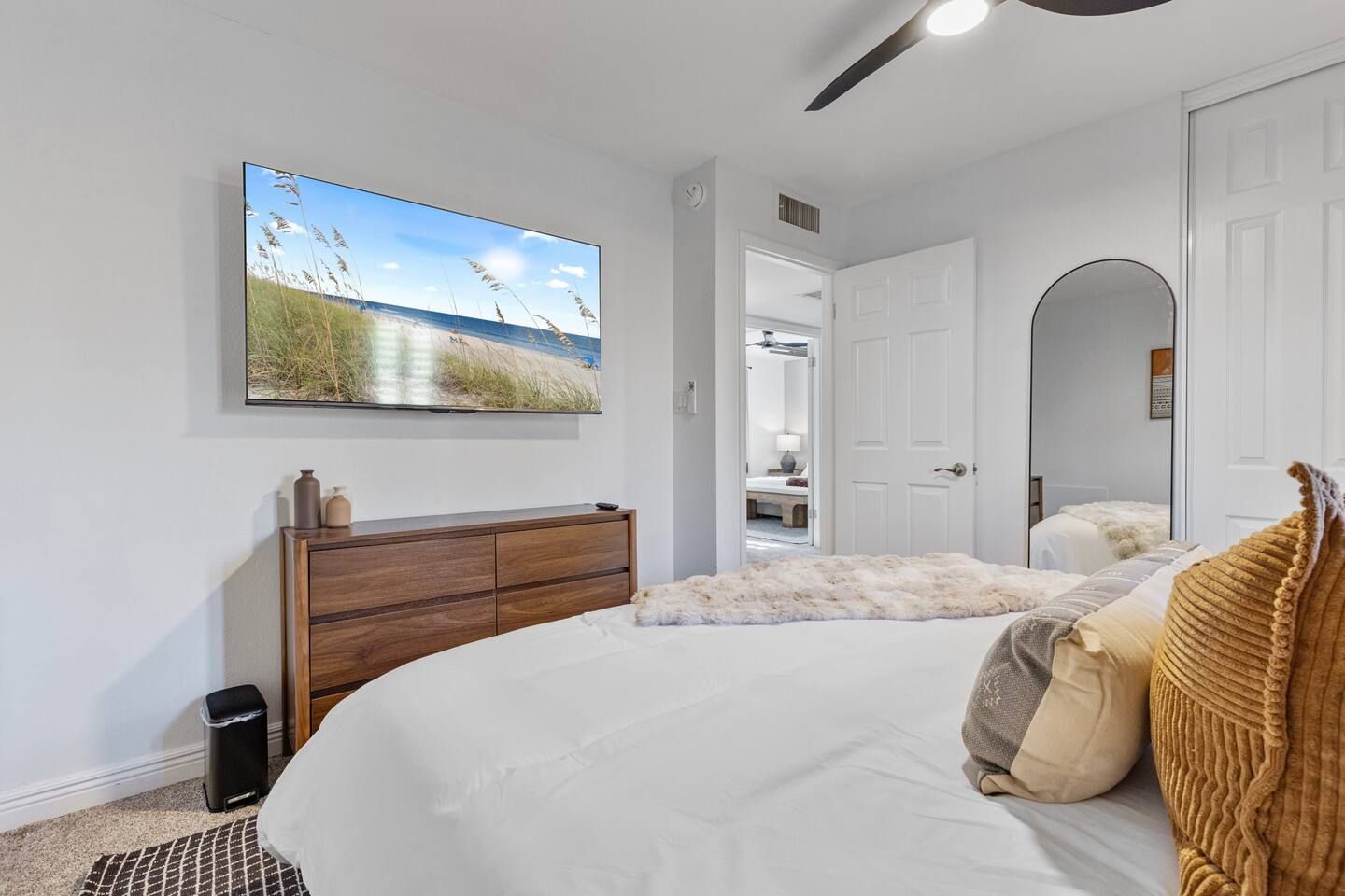 Une chambre avec un grand lit, une commode en bois, un téléviseur fixé au mur et une porte donnant sur une autre pièce.