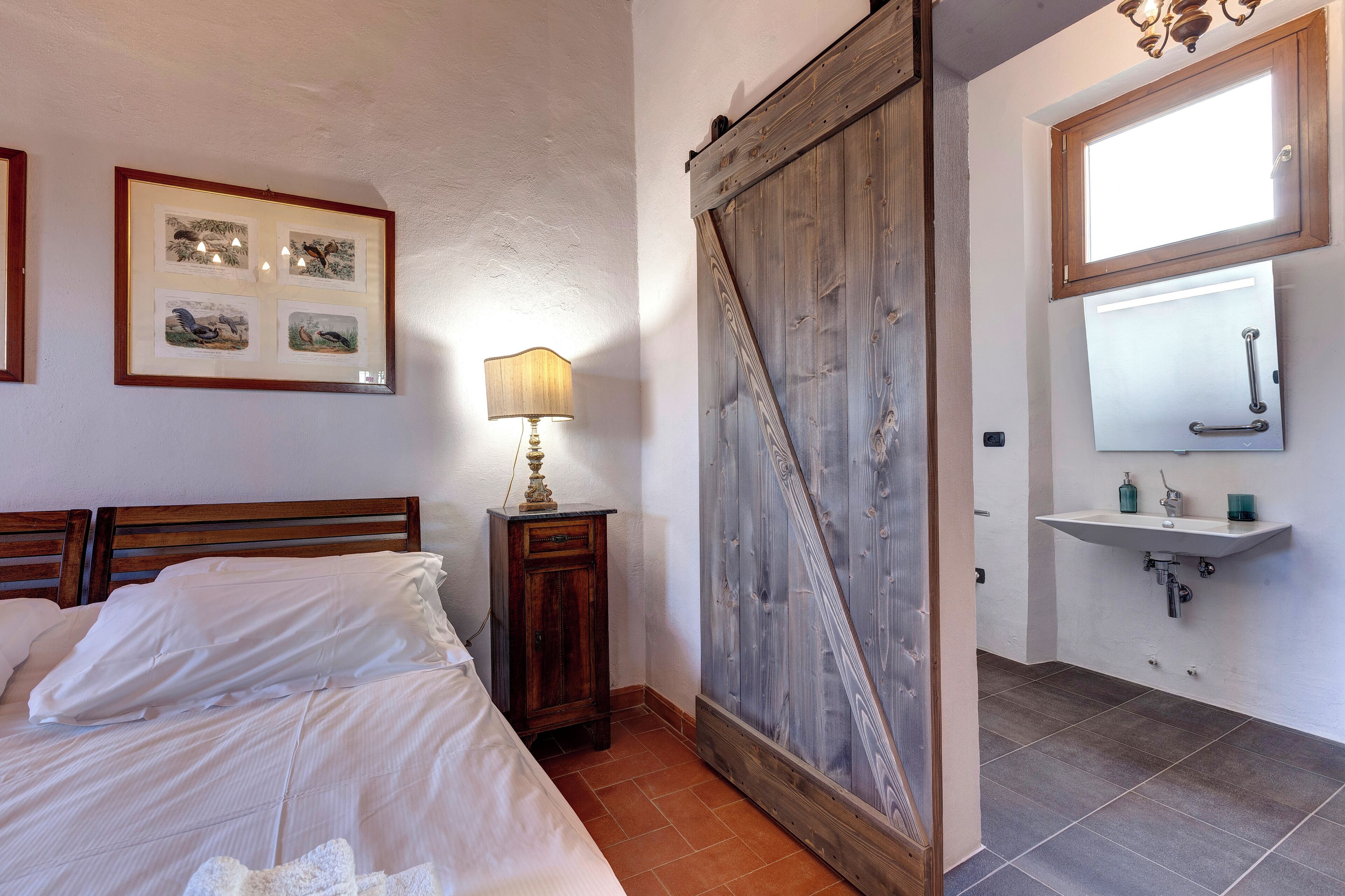 A bedroom with a bed, a nightstand with a lamp, a wooden wardrobe, and a bathroom with a sink and mirror.