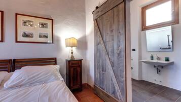 A bedroom with a bed, a nightstand with a lamp, a wooden wardrobe, and a bathroom with a sink and mirror.