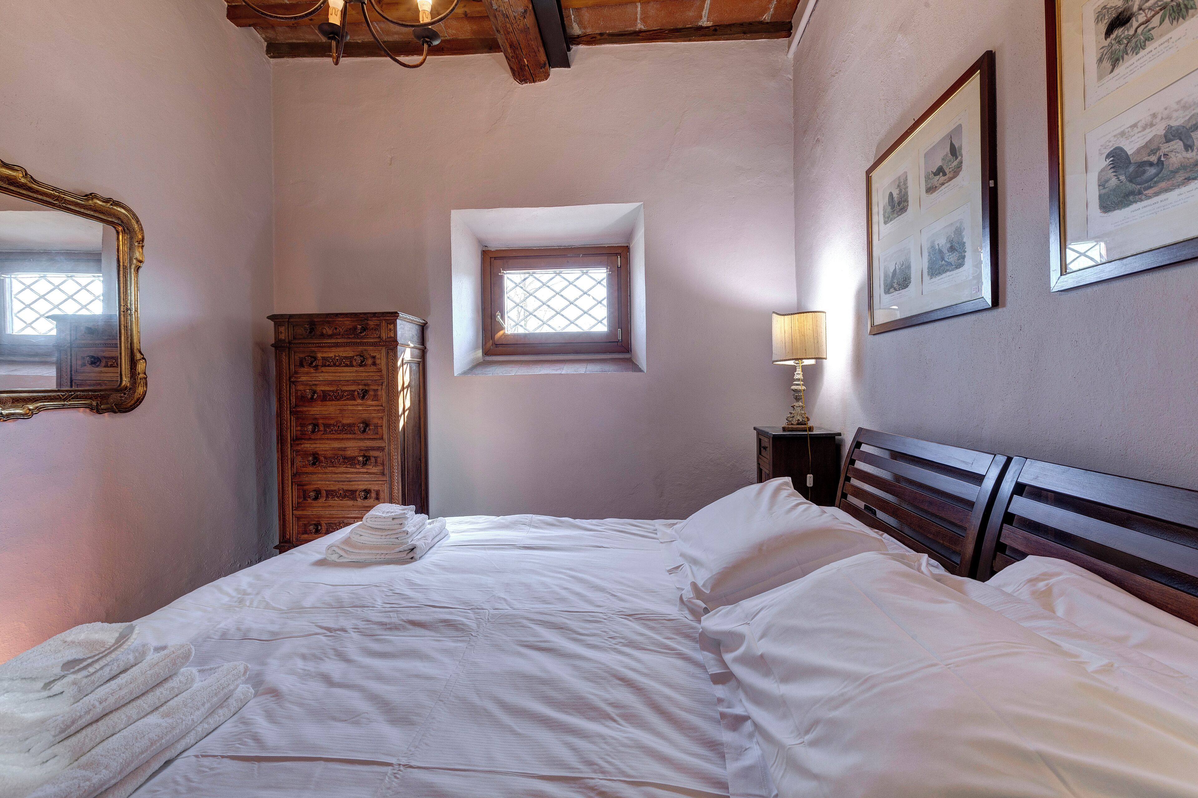 Un lit bien fait, avec du linge de lit blanc, une commode en bois dotée de tiroirs, une petite fenêtre et une lampe fixée au mur.