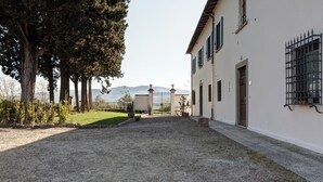 Un patio con un camino de grava que conduce a una entrada con reja, rodeado de árboles y un edificio de fachada blanca y marcos de ventana negros.