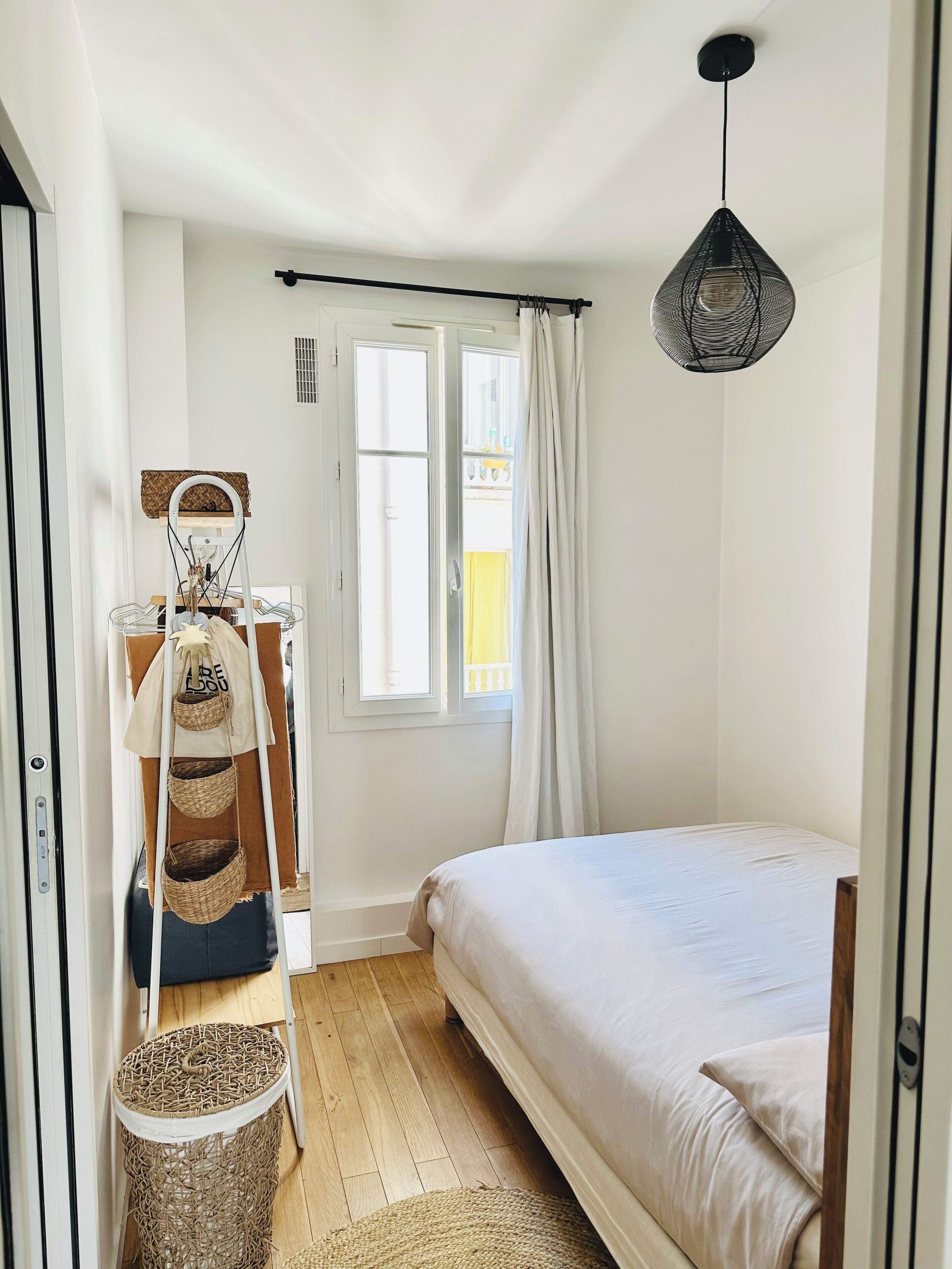 A bedroom with a bed, a hanging light fixture, a window with curtains, and a wooden floor.