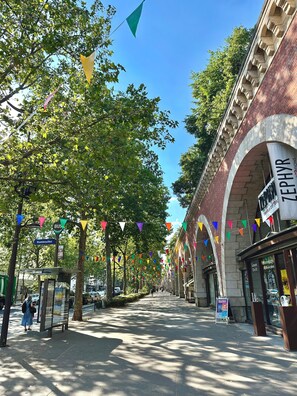 A street lined with trees and colorful triangular flags, a bus stop, and a building with an arched entrance. - Quiet & Bright • Line 1 • Direct access to the center (Paris)