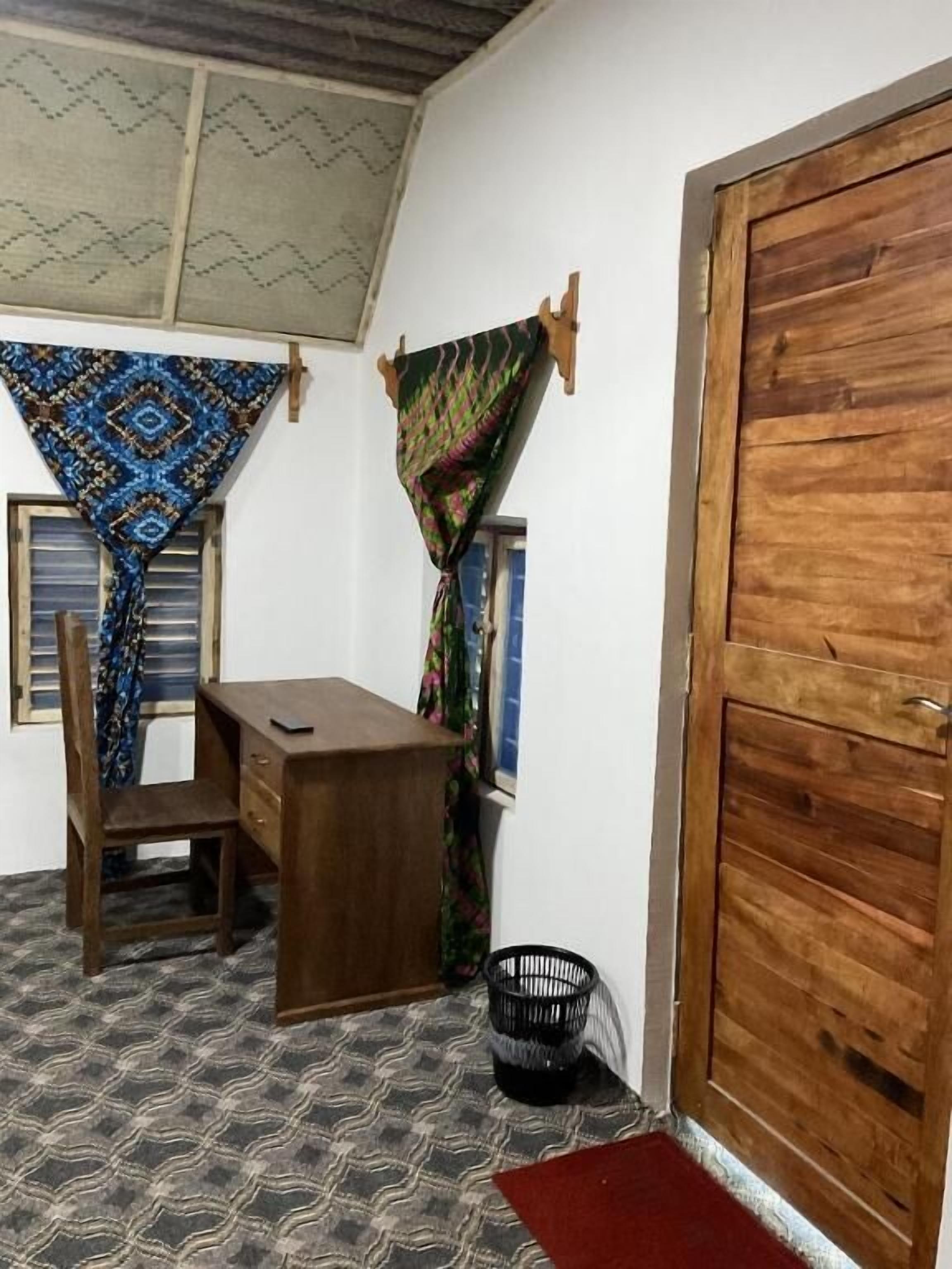 Una habitación con un escritorio de madera, una silla, un tapete estampado, una puerta de madera y una ventana con cortinas.
