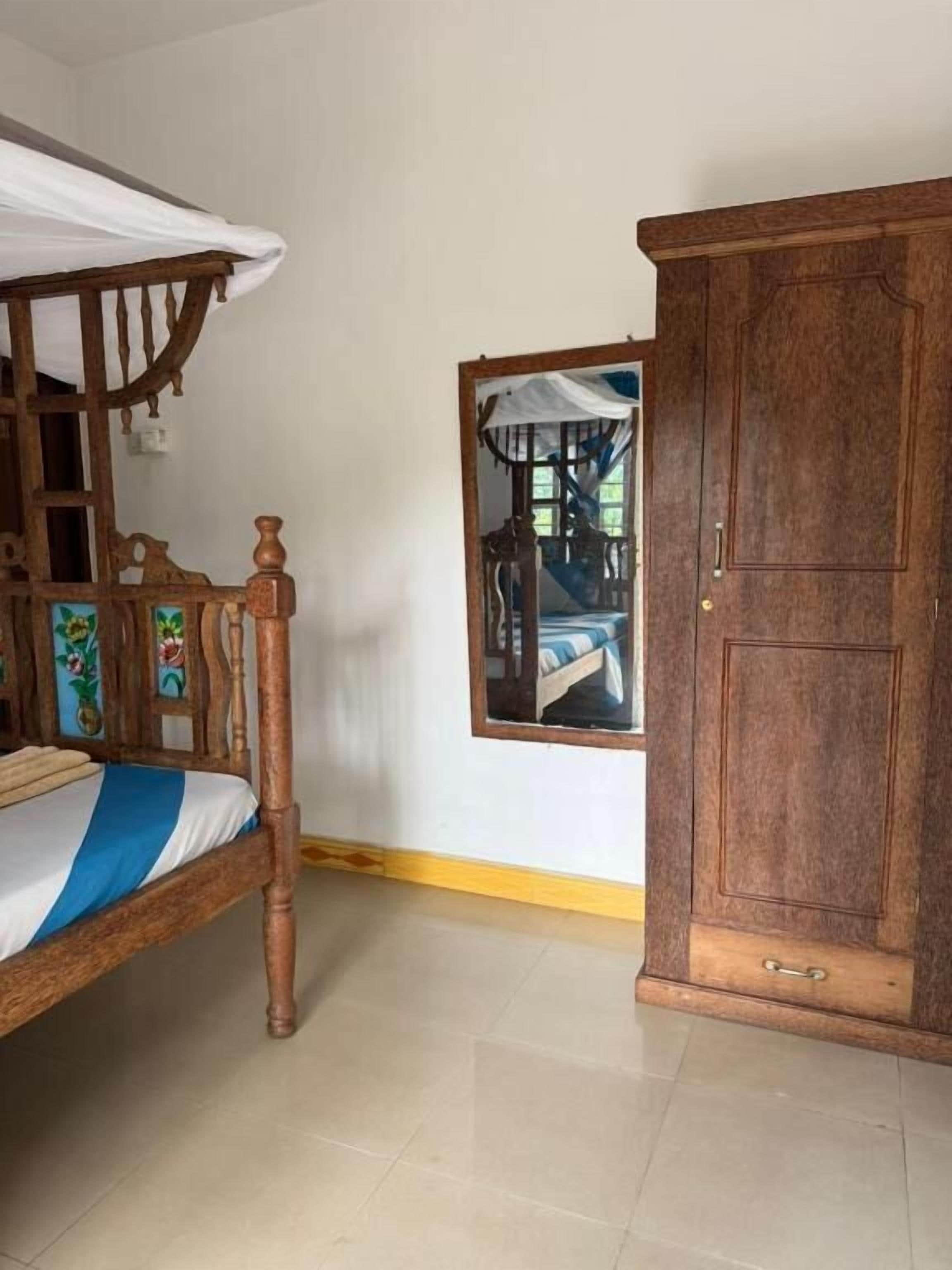 A room with a wooden bed, a wardrobe, and a mirror reflecting a bed with a blue bedspread.