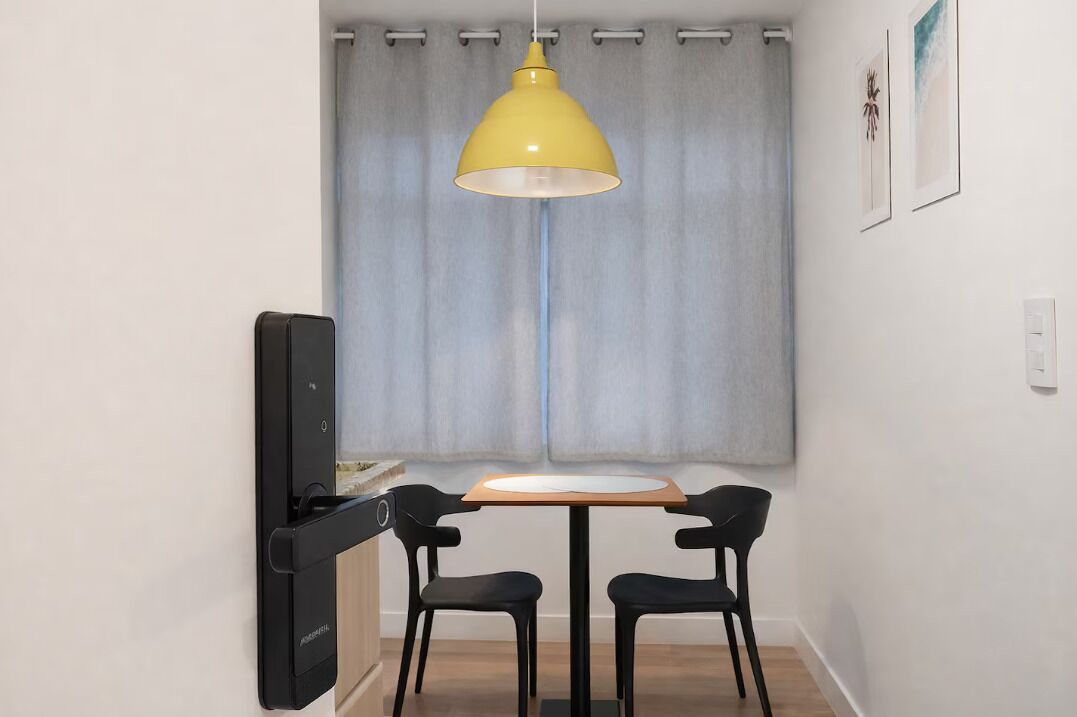 Un petit coin repas avec une table et deux chaises, un luminaire suspendu jaune, un téléviseur posé sur un meuble et une fenêtre avec des rideaux.