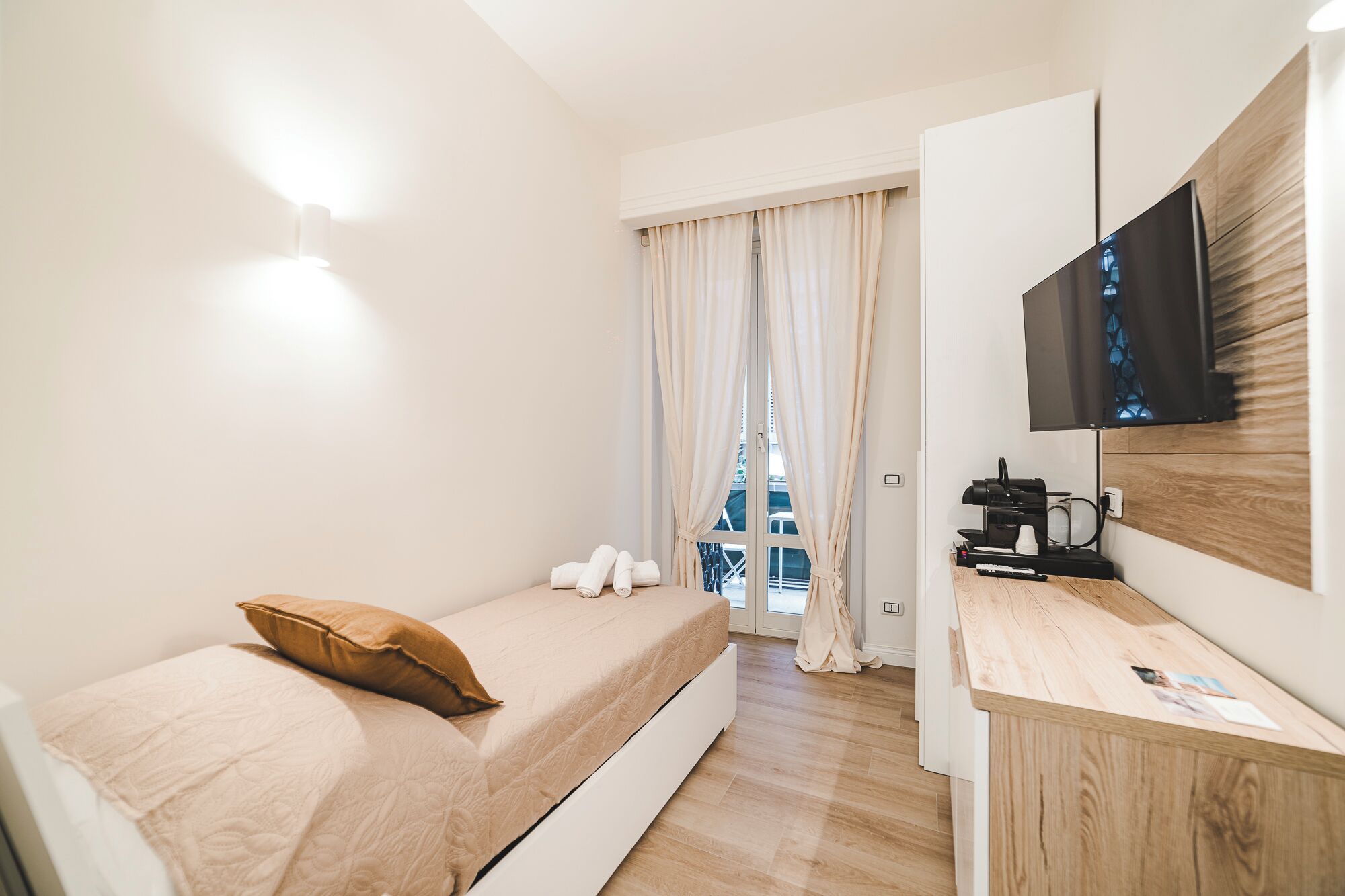 A hotel room with a bed, a desk with a coffee machine, a TV, and a window with curtains.
