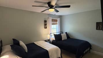 A bedroom with two beds, a ceiling fan, a window with curtains, and a bedside table with a lamp.