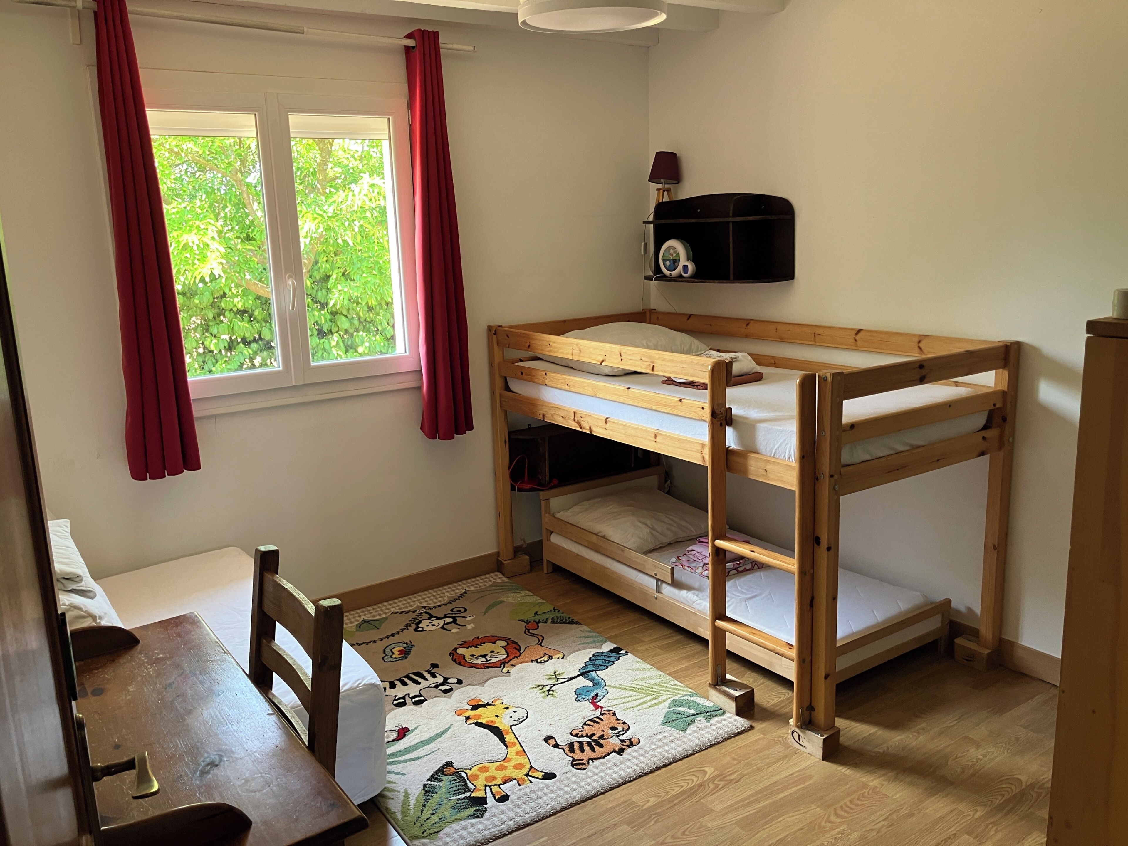 Quarto de criança com beliche, escrivaninha, cadeira, tapete com estampas de animais e janela com cortinas.