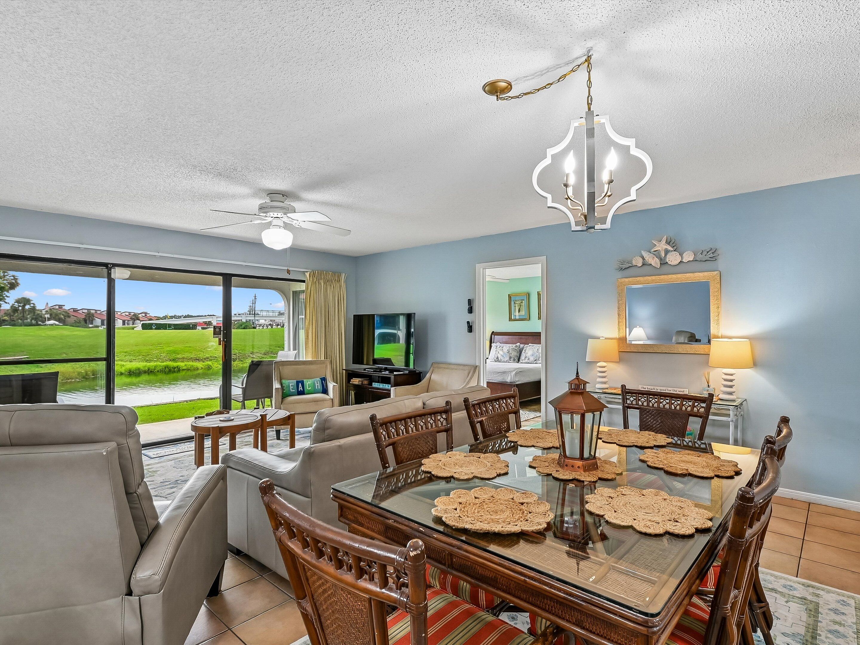 A spacious living room with a large glass door leading to a view of a golf course, a dining area with a glass table, and a bedroom visible through an open door.
