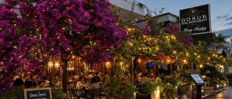 Un espacio al aire libre con mesas y sillas, rodeado de plantas florecientes e iluminación decorativa.