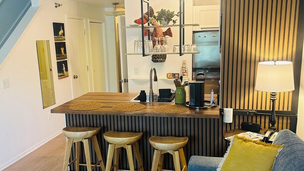A modern kitchen with a central island, bar stools, and a dining area with a sofa and cushions. - 2-bedroom 2-story renovated unit at The Cove Resort, park and walk downtown! (Lake Geneva)