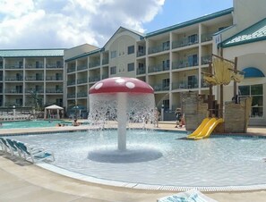 Un espace piscine d’hôtel agrémenté d’un élément aquatique en forme de champignon, d’un toboggan et d’un immeuble à plusieurs étages doté de balcons.