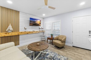 Ein modernes Wohnzimmer mit einer Wand in Holzoptik, einem Wand-Fernseher, einem Deckenventilator, einem beigefarbenen Sessel, einem kleinen runden Tisch und einem gemusterten Teppich.