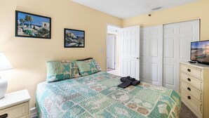A bedroom with a bed, a nightstand, a dresser, a television, and two doors. - Beach Front 3 Bedroom - Beach Place #507 (Madeira Beach)
