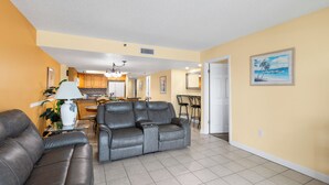 A living room with a gray leather sofa, a dining area with a table and chairs, and a kitchen in the background. - Beach Front 3 Bedroom - Beach Place #507 (Madeira Beach)
