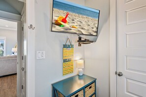 A television mounted on a wall with a beach scene, a 'BEACH Rules' poster, a wall-mounted lamp, and a small table with baskets. - Easy Breezy! 4 bedroom 4 Bath, Direct Ocean view, easy beach access. (Crystal Beach)