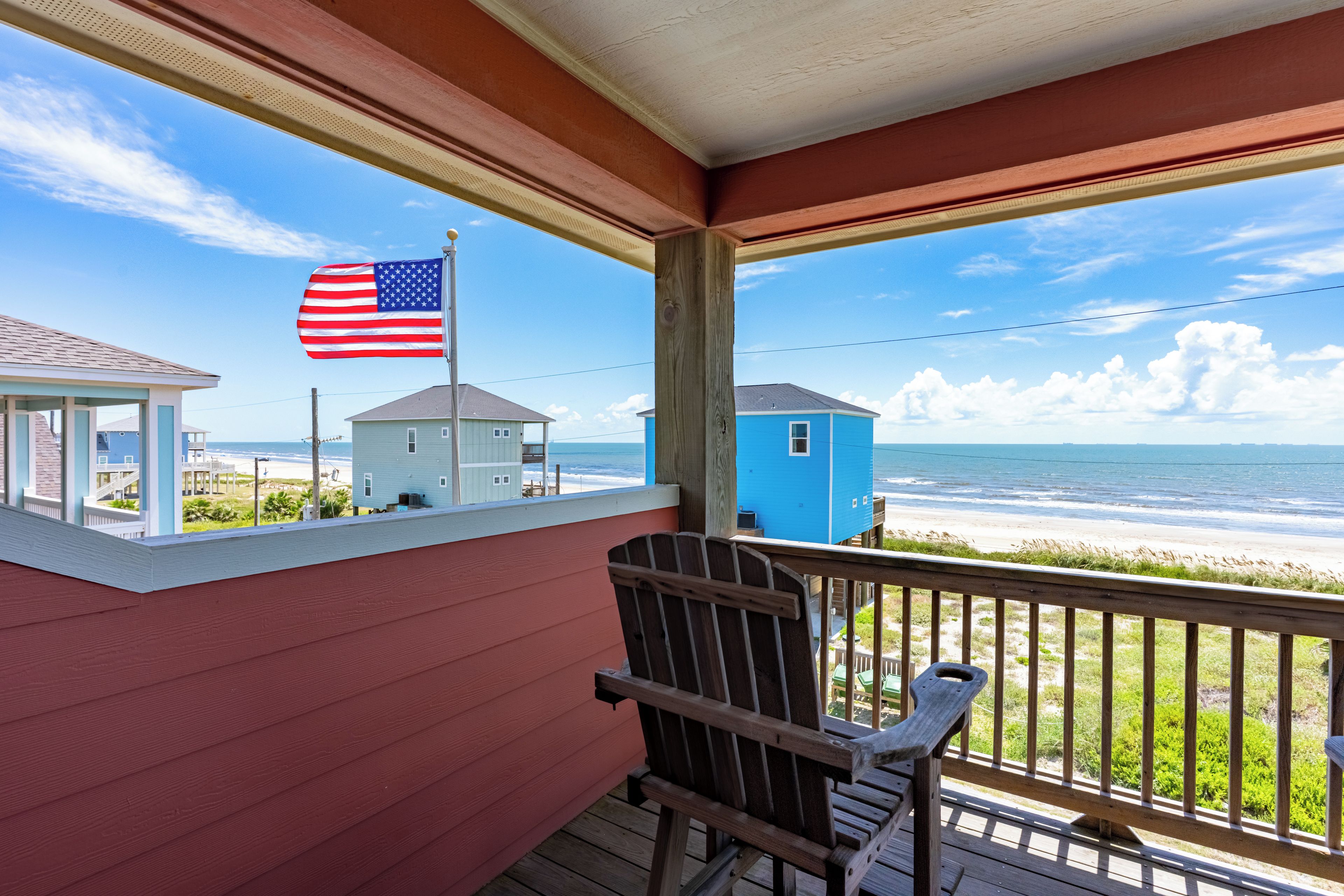 A wooden deck with a view of the beach and ocean, a flag, and a small blue building.