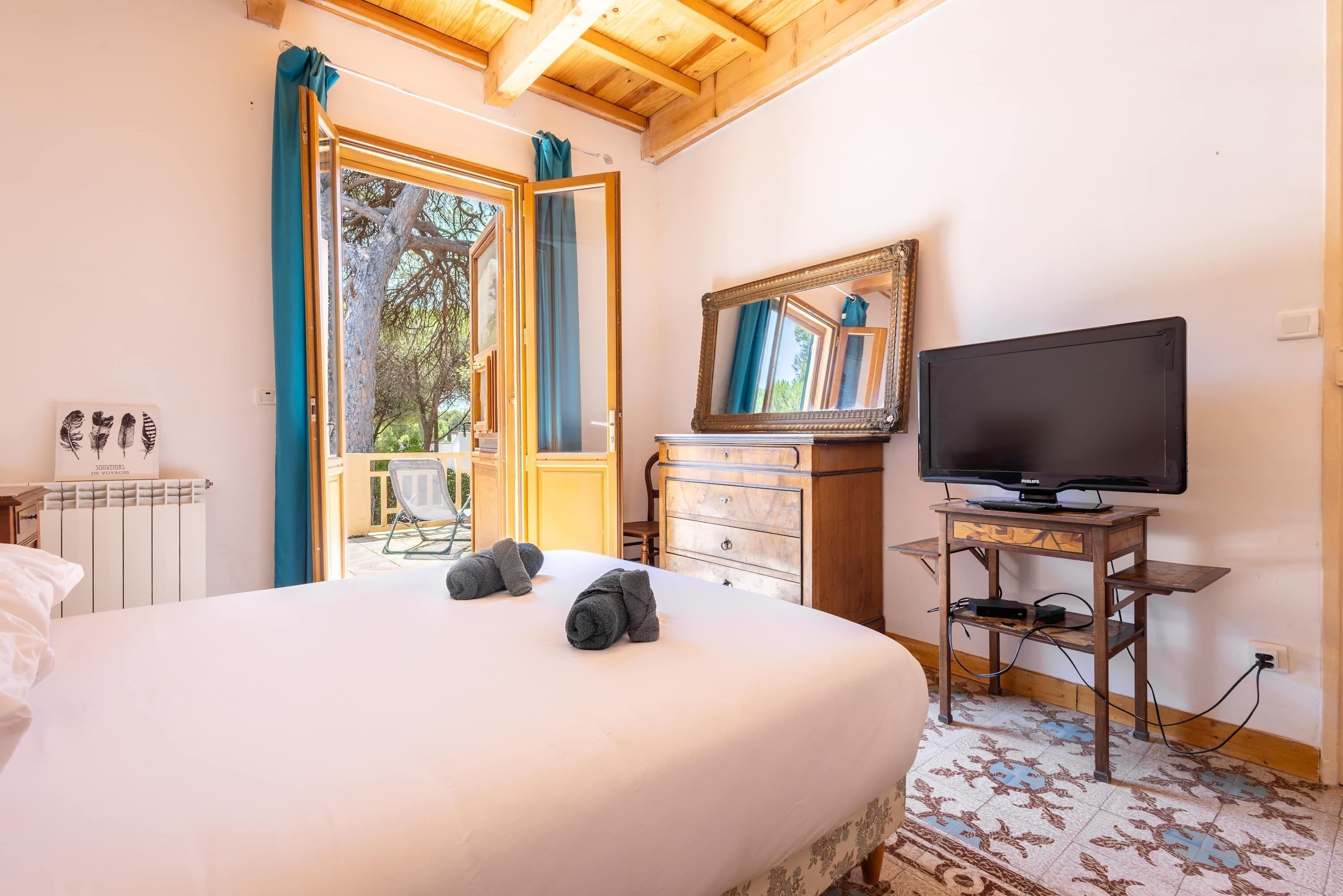 A bedroom with a bed, a television on a wooden stand, a dresser, and a window with curtains.